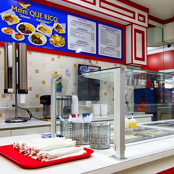 A counter at MMM Que Rico with a menu board, food warmers, and wrapped utensils.