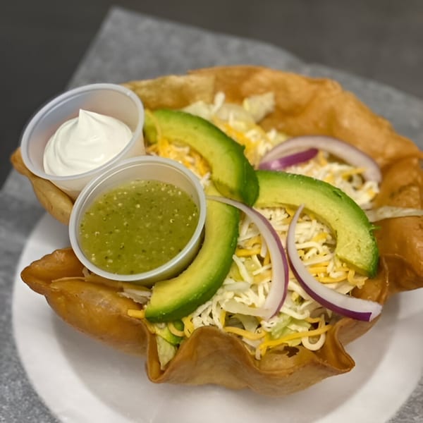 A taco salad in a crispy shell with avocado, red onion, cheese, and two dipping sauces.