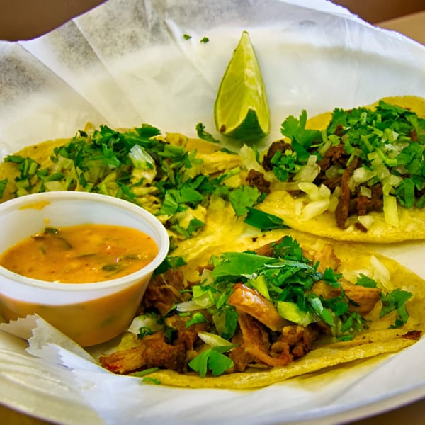 Three Latin-American tacos with meat, cilantro, and onions, served with a side of orange sauce and lime.