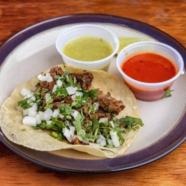 A MMM Que Rico taco with meat, cilantro, and onions, served with green and red sauces.