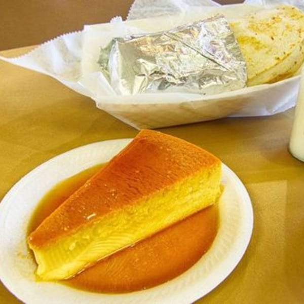 A slice of flan with caramel sauce on a white plate, with a wrapped item in the background.