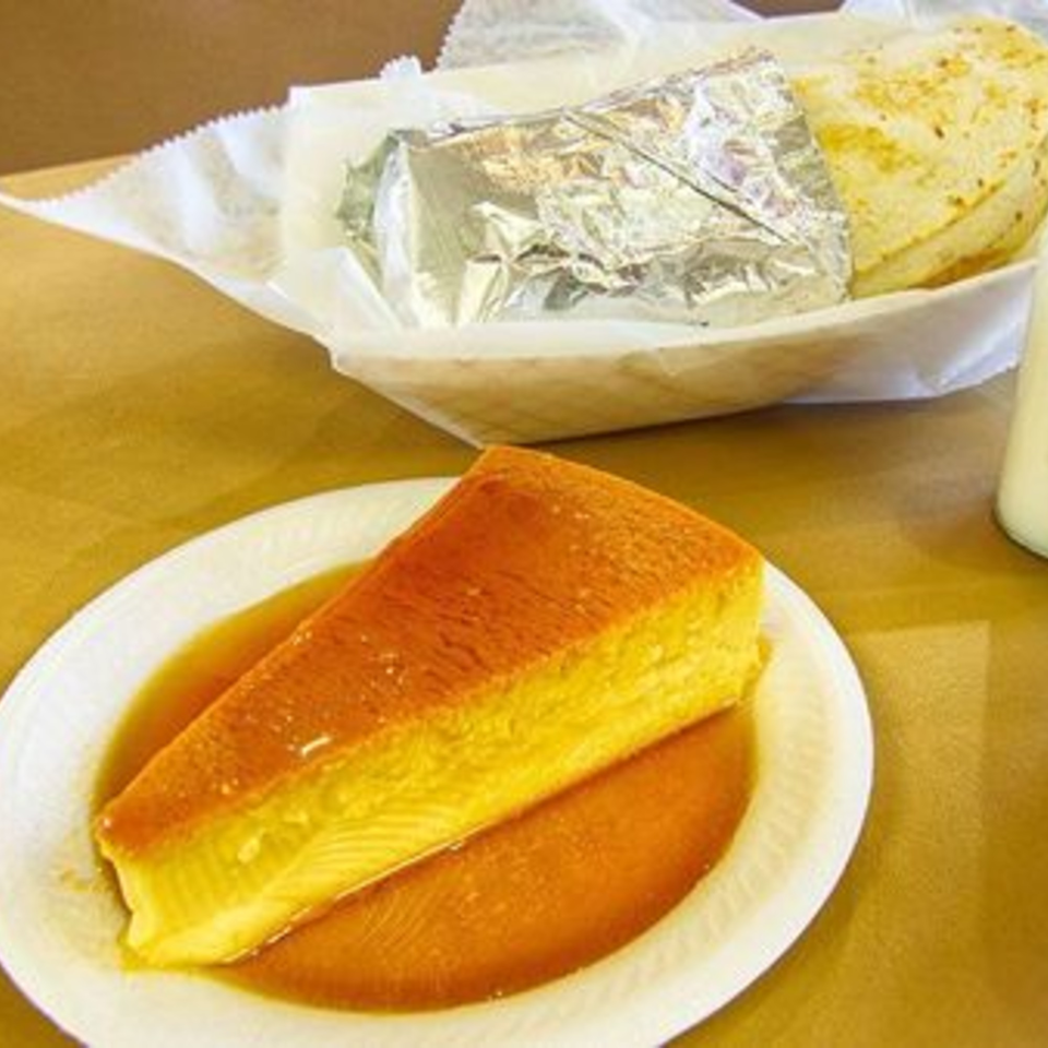 A slice of flan with caramel sauce on a white plate, with a wrapped item in the background.