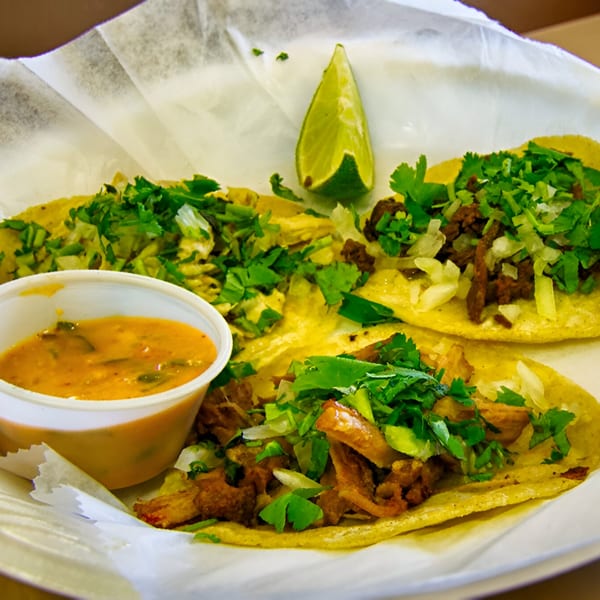 Three MMM Que Rico tacos with meat, cilantro, onions, a side of sauce, and a lime wedge.